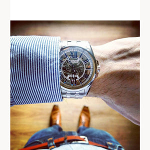 Close-up of a wrist wearing a silver watch with a detailed face, dressed in a striped shirt and jeans.