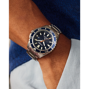 Wrist wearing a silver and blue watch with a brand logo on a neutral background