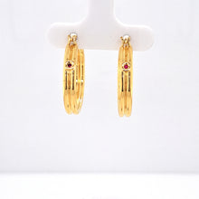 Load image into Gallery viewer, A pair of gold hoop earrings with a small round red gemstone set on each hoop, displayed on a white jewelry stand against a white background.
