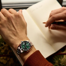 Load image into Gallery viewer, Person wearing a watch with a green face, sitting at a desk with a notebook and pen.
